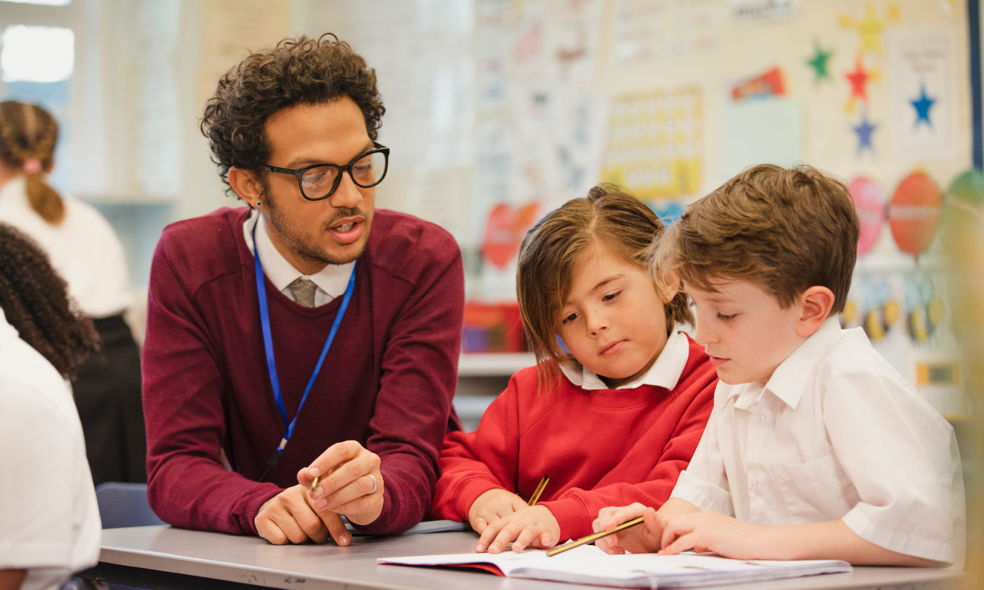 A teacher supporting two primary school pupils as they work together on a classroom activity at a desk. The teacher is wearing glasses, a burgundy jumper and a blue lanyard, while the children are writing in an exercise book. Colourful classroom displays are visible in the background.